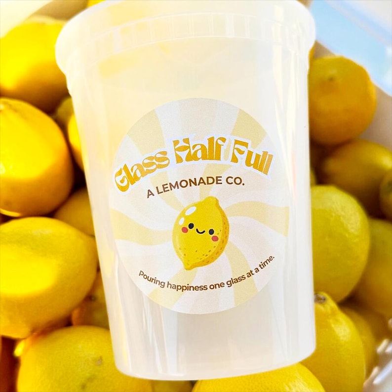 May include: A clear plastic cup featuring a circular sticker with a yellow and white sunburst pattern. The text reads Glass Half Full A LEMONADE CO. and Pouring happiness one glass at a time, with a central illustration of a smiling yellow lemon.