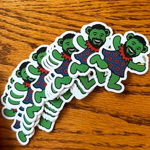 May include: A stack of die-cut stickers featuring a green bear character with a black beard and hair. The bear wears a blue dress with red circular patterns and a red collar, and is holding a small lit cigarette in one hand.