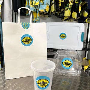 May include: A white paper shopping bag, a white plastic container, a clear plastic container, and a clear plastic cup, all with the logo "The Lemon Wagon" on them. The logo is a blue circle with yellow text.