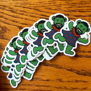 May include: A stack of die-cut stickers featuring a green bear character with a black beard and hair. The bear wears a blue dress with red circular patterns and a red collar, and is holding a small lit cigarette in one hand.