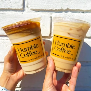May include: Two clear plastic cups filled with iced coffee beverages. Each cup features a yellow rectangular label with the black text Humble Coffee and the phrase It is what it is. The left cup contains a layered drink with dark brown foam on top.