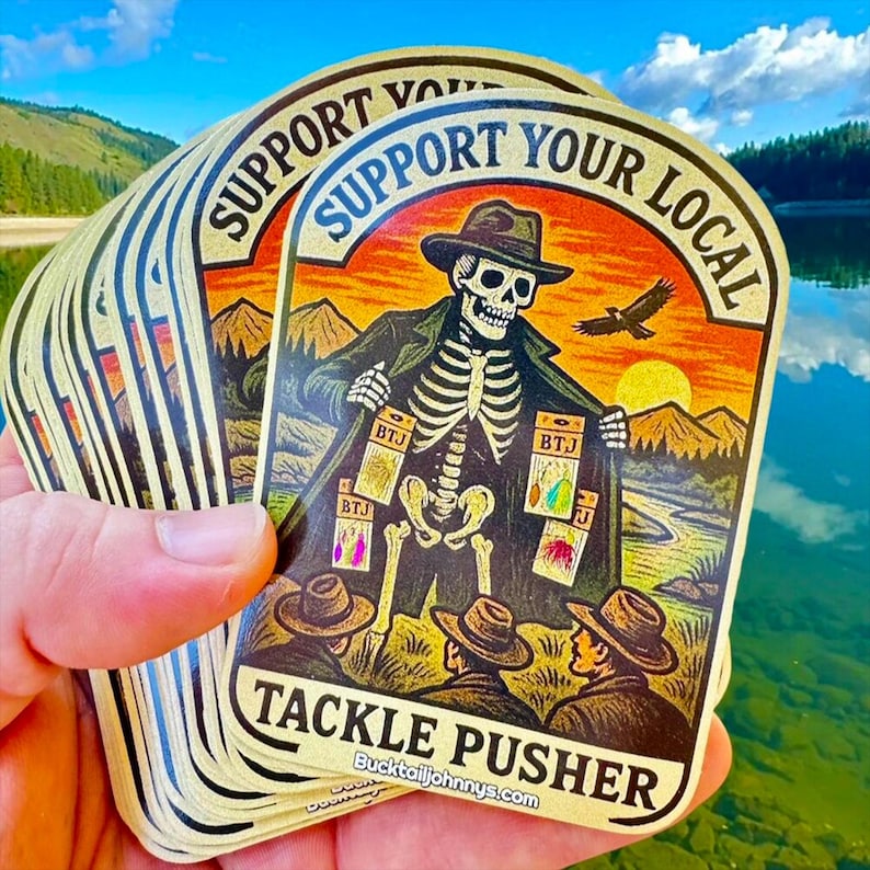 May include: A stack of die-cut stickers featuring a skeleton wearing a hat and coat, opening the coat to reveal fishing lures. The text reads SUPPORT YOUR LOCAL TACKLE PUSHER and bucktailjohnnys.com. The background shows mountains and a bird.