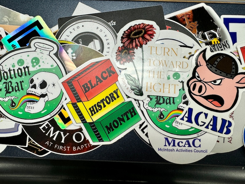 May include: A collection of assorted vinyl stickers featuring various designs including a skull with a rainbow, a Black History Month graphic, a pig wearing a helmet with the text AGAB, and a floral illustration with the text TURN TOWARD THE LIGHT.