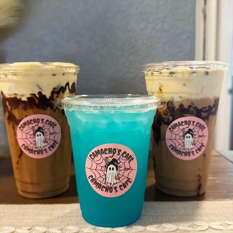 May include: Three clear plastic cups with lids. Two cups contain a brown iced coffee beverage with dark chocolate drizzle and a light-colored foam top. One central cup contains a bright blue liquid. Each cup features a round pink sticker with a ghost graphic and the text Camacho's Cafe.