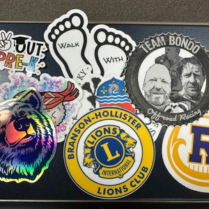 May include: A collection of colorful stickers with various designs. Notable stickers include a holographic bear face, a Lions Club logo, a sticker with the text "OUT PRE-K", and a sticker with the text "WALK WITH KY".