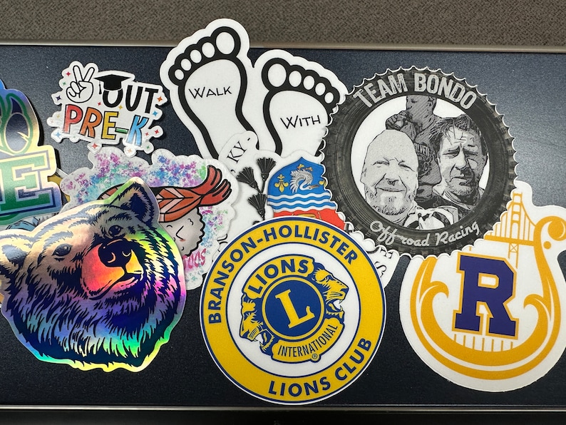 May include: A collection of colorful stickers with various designs. Notable stickers include a holographic bear face, a Lions Club logo, a sticker with the text "OUT PRE-K", and a sticker with the text "WALK WITH KY".