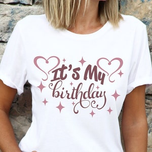 May include: White t-shirt with the text "It's My Birthday" in pink script with hearts and stars around the text.