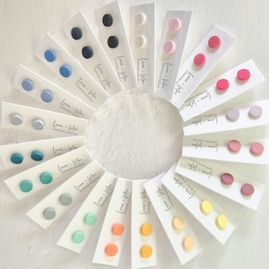 May include: A selection of handmade stud earrings in a range of colours, such as blue, pink, and yellow, displayed in a circular arrangement. Each pair is presented on a white card with the text "Luna Mae Handmade Jewellery".
