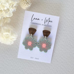 May include: A pair of teal blue flower-shaped earrings with pink centres and wood accents. The earrings are displayed on a white card with the text "Luna + Mae Handmade Jewellery Est. 2022".