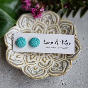 Op de afbeelding: Een paar tealblauwe stoffen oorbel studs met een wit geometrisch patroon. De oorbellen worden getoond op een witte kaart met de tekst "Luna & Mae Handmade Jewellery".