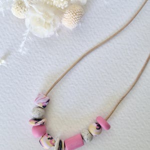 May include: A pink and white patterned necklace with a variety of beads, including round, square, and rectangular shapes. The necklace is made with a brown leather cord.