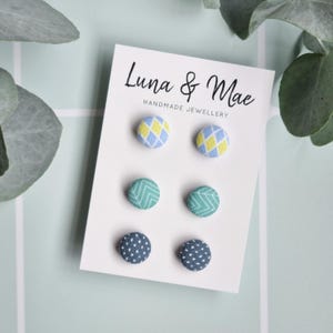 Op de afbeelding: Een set van zes stoffen knoop oorbellen op een witte kaart met de tekst "Luna & Mae Handmade Jewellery". De oorbellen zijn in drie verschillende patronen: blauwe en gele ruiten, groene en witte chevron en blauwe en witte stippen.