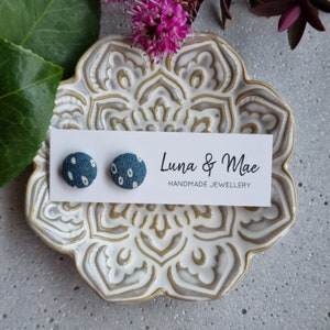 Puede incluir: Un par de pendientes de botón de tela azul y blanca con estampado de lunares. Los pendientes se muestran en una tarjeta blanca con el texto "Luna & Mae Handmade Jewellery".