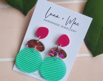 May include: A pair of handmade earrings featuring a layered design with a green striped circle, a pink circle, and a multi-colored glitter circle. The earrings are displayed on a white card with the text "Luna + Mae Handmade Jewellery Est. 202..."