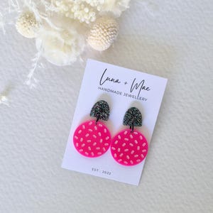 Puede incluir: Un par de pendientes redondos de color rosa y negro con un diseño moteado. Los pendientes están hechos de arcilla polimérica y están sobre una tarjeta blanca con el texto "Luna + Mae Handmade Jewellery Est. 2022".