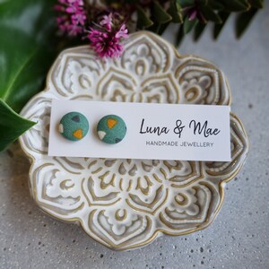 May include: A pair of green fabric stud earrings with a geometric pattern of white and yellow triangles. The earrings are displayed on a white card with the text "Luna & Mae Handmade Jewellery".