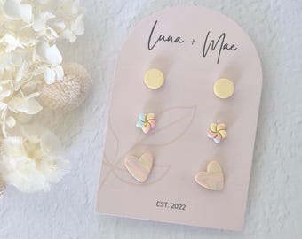 Puede incluir: Un conjunto de pendientes Luna + Mae exhibidos en una tarjeta rosa. Los pendientes incluyen dos pares de tachuelas amarillas, dos pares de pendientes en forma de flor pastel y dos pares de pendientes en forma de corazón. La tarjeta dice "Luna + Mae" y "EST. 2022".