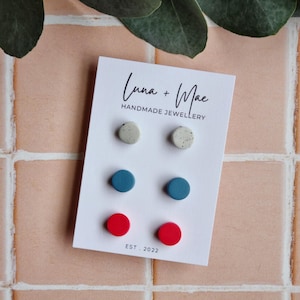 May include: A set of six small, round, stud earrings. The earrings are made of clay and come in three colors: white with black speckles, blue, and red. The earrings are displayed on a white card with the text "Luna + Mae Handmade Jewellery" and "Est. 2022".