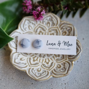 Puede incluir: Un par de pequeños pendientes redondos de tela azul con un patrón floral blanco. Los pendientes se muestran en una tarjeta blanca con el texto "Luna & Mae Handmade Jewellery".