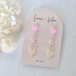 以下が含まれることがあります： カラフルなポリマークレイ製のイヤリング6個セット。セットにはピンクの円形が2つ、黄色とピンクの花の形が2つ、黄色とピンクのハートの形が2つ含まれています。イヤリングは「Luna + Mae Est. 2022」というテキストが書かれたカードに飾られています。