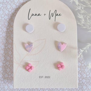 May include: A cream-coloured display card featuring four pairs of earrings. The earrings include white circles, marbled hearts, and pink flower shapes. The card is printed with the text "Luna + Mae" and "EST. 2022".