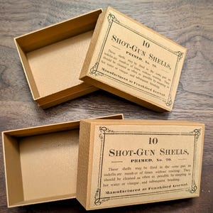 May include: Two vintage Shot-Gun Shells boxes, open and closed, with text. The boxes are tan with black text and decorative borders. The text reads "10 Shot-Gun Shells, Primed, No. 20." The boxes are made of cardboard.