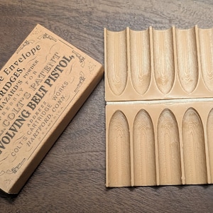 May include: A vintage cartridge box and a wooden bullet holder. The box is tan with black text that reads "6 Combustible Envelope Cartridges for Colt's Revolving Belt Pistol." The bullet holder is light brown and has 12 bullet slots.