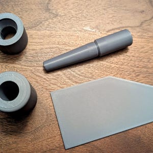 May include: Two dark gray cylindrical components with hollow centers, a dark gray tapered rod-like piece, and a flat, light gray trapezoidal plate with a textured surface finish, all arranged together as a set of separate 3D printed parts.