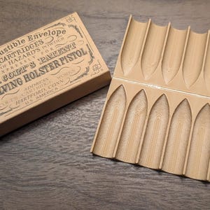 May include: A vintage-style box and a wooden cartridge holder. The box is tan with black text that reads "Combustible Envelope Cartridges" and "Col. Colt's Patent Revolving Holster Pistol." The holder has 12 slots, each approximately 1.5 inches long.
