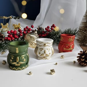 May include: Three ceramic nutcracker-themed containers in green, white, and red, with gold accents. The containers are arranged on a white surface with Christmas decorations, including red berries, pine needles, and gold ornaments.