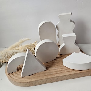 May include: A set of seven white geometric shapes made of plaster or ceramic. The shapes include a circle, a half-circle, a triangle, a rectangle, and three abstract shapes. The shapes are arranged on a wooden tray.