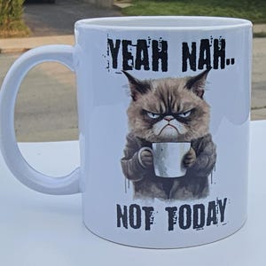 May include: White ceramic coffee mug featuring a grumpy cat cartoon holding a coffee mug. The text on the mug reads "Yeah Nah... Not Today."