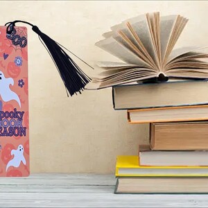Puede incluir: Un marcapáginas rosa y naranja con un fantasma y el texto "Spooky Book Season". El marcapáginas tiene un borla negra.