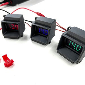 May include: Three black digital voltmeters with red, blue, and green displays. The voltmeters show readings of 13.9, 14.0, and 14.0. Two red wire connectors are also visible.