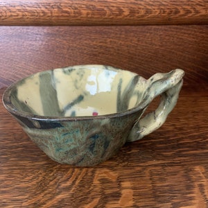 May include: A handmade ceramic bowl with a unique handle. The bowl features a blend of green, blue, and cream colors with a textured surface. The handle is organically shaped, adding to the rustic aesthetic. The bowl is suitable for serving soups or other dishes.