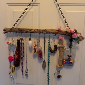 May include: A rustic wall hanging crafted from a natural branch, suspended by a chain. It features an assortment of decorative items, including small bottles, beads, and a spoon. Pink roses and other embellishments add a touch of color and charm to this unique home decor piece.