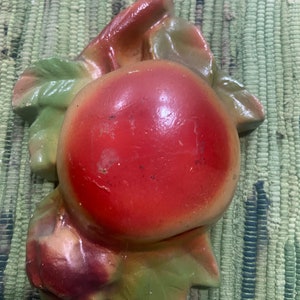 May include: A vintage, hand-painted ceramic apple wall hanging. The apple is a vibrant red with subtle shading, and is surrounded by green and brown leaves. The piece has a glossy finish and is likely from the mid-20th century.