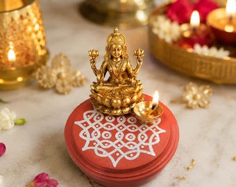 May include: A gold-toned metal figurine of the deity Lakshmi seated on a lotus flower, placed atop a circular red base decorated with a white geometric pattern. A small brass oil lamp with a lit flame sits beside the figurine on the red base.