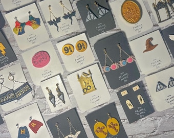 May include: A collection of handmade earrings on display, featuring various designs such as lightning bolts, bats, and Hogwarts-themed elements. Each pair is attached to a card with the text "PAJAMA MAMA Co."