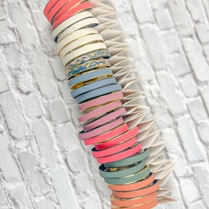 May include: A display of colorful, stacked bracelets. The bracelets feature a gold-colored band with various shades of pink, coral, blue, and green leather. The bracelets are stacked on a white, fan-shaped display against a white brick wall.