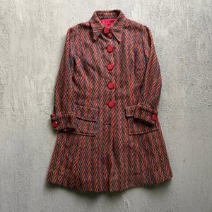 May include: A long-sleeved coat featuring a vertical herringbone pattern in shades of orange, blue, and grey. The coat has a pointed collar, a front button closure with five large red buttons, two front flap pockets, and red buttons on the cuffs.