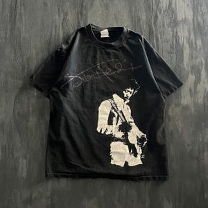 May include: A black short-sleeved t-shirt featuring a white graphic print of Jimi Hendrix playing an electric guitar. Above the figure is a white stylised signature. The shirt has a crew neckline and a standard fit, made of cotton fabric.