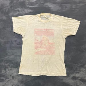 May include: A cream-colored short-sleeve t-shirt featuring a faded red graphic print on the chest. The graphic depicts a building and includes the text Grateful Dead and Live at the Great American Music Hall. The shirt has a crew neckline.