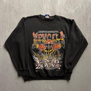 May include: A black long-sleeve crewneck sweatshirt featuring a graphic print with the text CINCINNATI BENGALS END ZONE. The design includes three orange and black football helmets with yellow lightning bolts and white smoke accents.