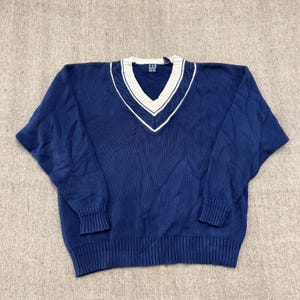 Könnte beinhalten: Ein marineblauer Langarmpullover mit V-Ausschnitt und weißem Zopfstrickbesatz. Der Pullover hat gerippte Bündchen an Ärmeln und Saum. Das Etikett im Nackenbereich zeigt den Text GAP in weißen Buchstaben auf dunkelblauem Hintergrund.