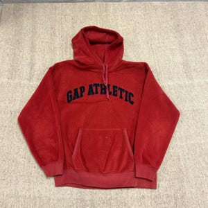 May include: A red fleece hooded sweatshirt featuring the text GAP ATHLETIC in dark blue lettering across the chest. The garment includes a front kangaroo pocket, long sleeves with ribbed cuffs, a matching ribbed hem, and a drawstring hood.
