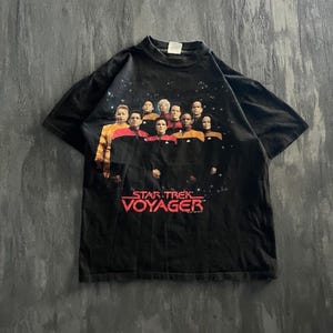 May include: A black short-sleeve t-shirt featuring a graphic print of the Star Trek Voyager cast in their uniforms against a starry space background. The red text Star Trek Voyager is printed below the cast members on the front of the garment.