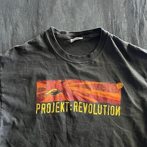 May include: A charcoal grey crew neck t-shirt featuring a graphic print of a red and orange stylised plug with radiating circular patterns. Below the graphic, the text PROJEKT: REVOLUTION is printed in bold yellow capital letters on the front.