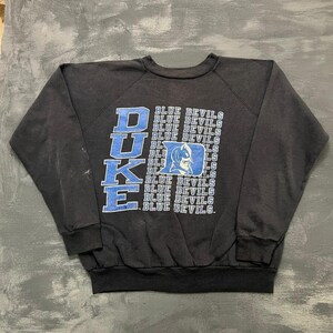 Pode incluir: Uma camisola preta de manga comprida com a palavra DUKE em letras azuis grandes no lado esquerdo e o texto BLUE DEVILS repetido num padrão de grelha à direita, centrado com um logótipo gráfico azul e branco da mascote Duke Blue Devils.