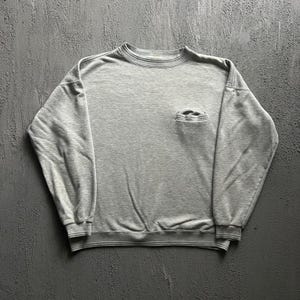May include: A light grey crewneck sweatshirt featuring long sleeves, ribbed cuffs, a ribbed hem, and a single small patch pocket located on the upper left chest area. The garment is constructed from a soft, heathered fabric with a standard fit.